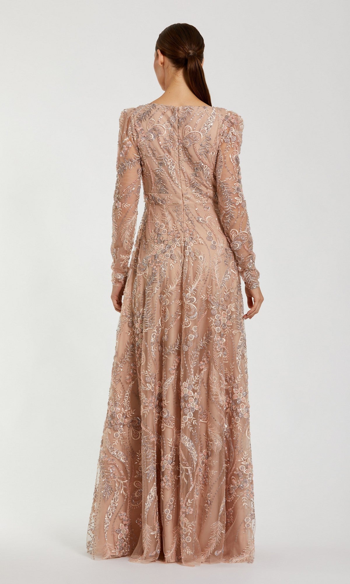 A woman stands facing away, wearing the Mac Duggal 68908 long formal dress—a floor-length, long-sleeved gown in blush pink with intricate silver embroidery on sheer fabric. Her brown hair is styled in a low half ponytail.